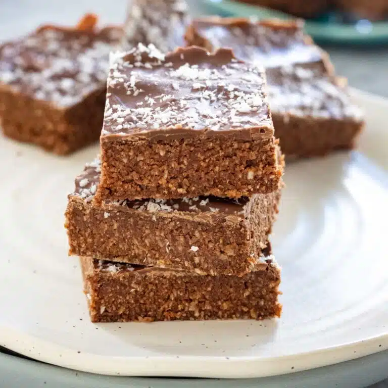 A square image of 3 pieces of coconut chocolate slice stacked on a white plate. Several more pieces are behind them on the same plate.