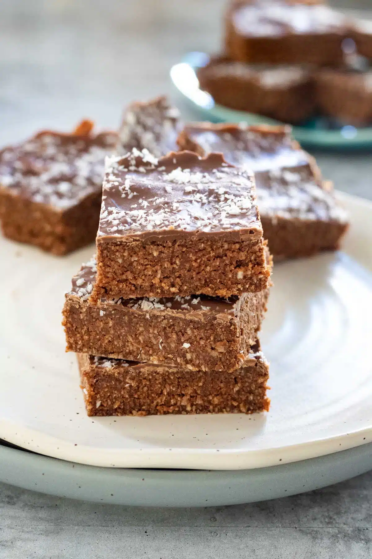 A stack of 3 coconut chocolate slice pieces are sitting on a white plate with a few behind them on the same plate. A small teal plate is also behind and to the right filled with more slice pieces.