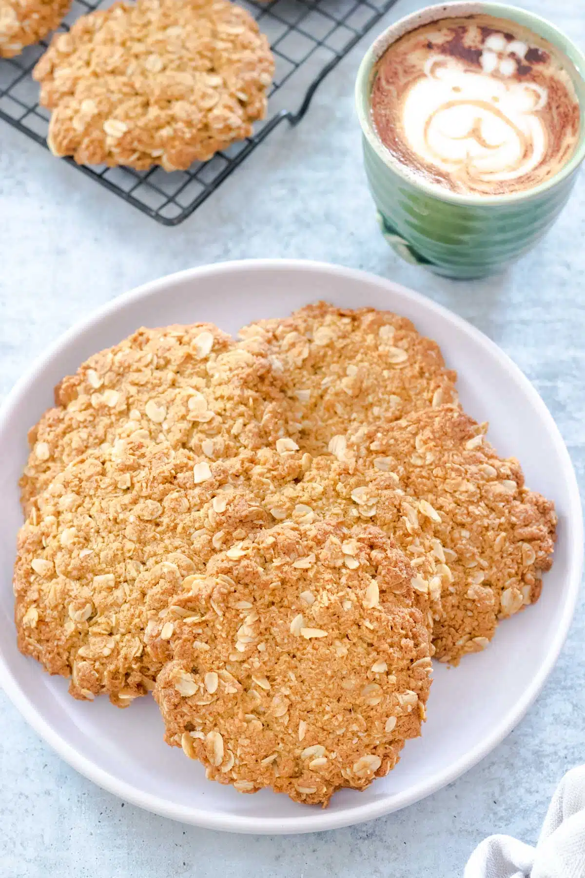 A light purple plate has been filled with golden Anzac Biscuits. A cup of coffee is in the background with a bear in latte art on the top. Just beside this is a cooling rack filled with more Anzac biscuits.