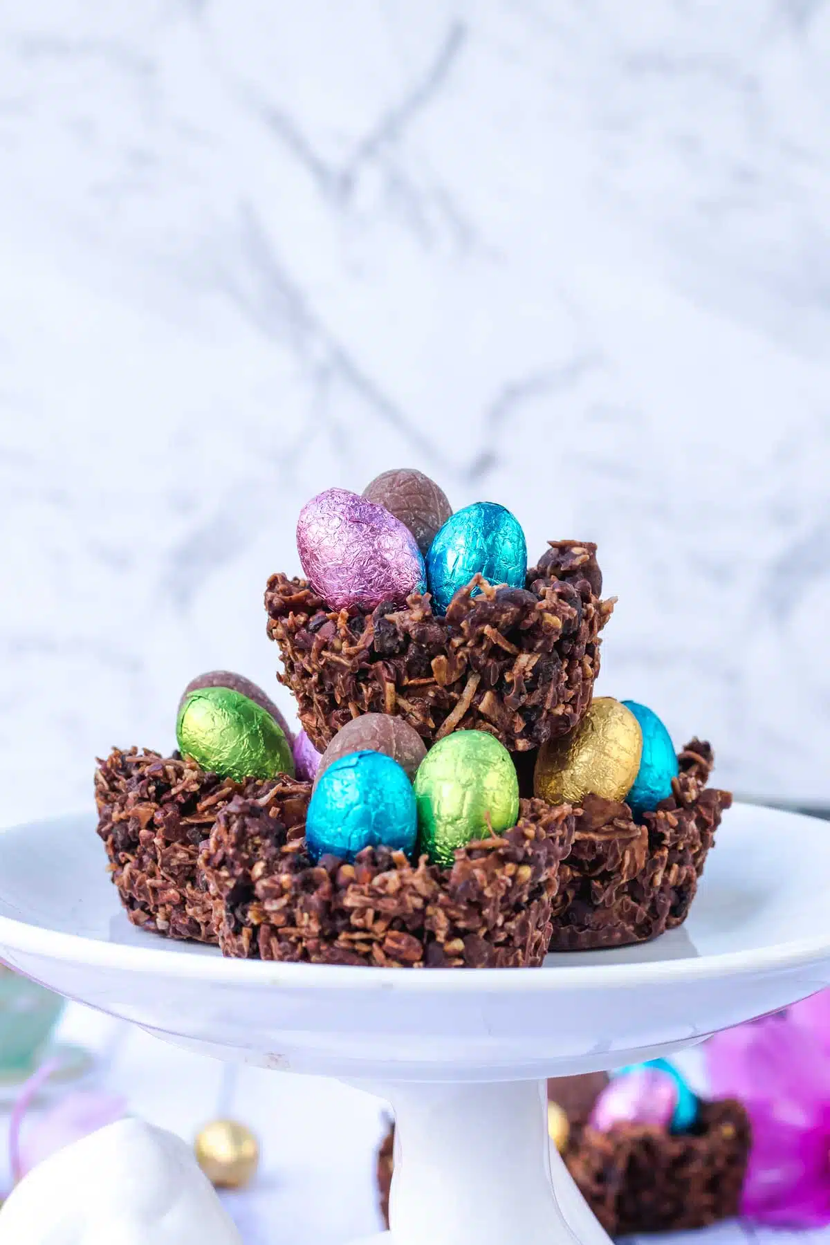 Three chocolate Easter nests have been piled onto a platter, which is a white rabbit holding a plate up on its feet. Other chocolate Easter nests can be seen on the table surrounding the rabbit.