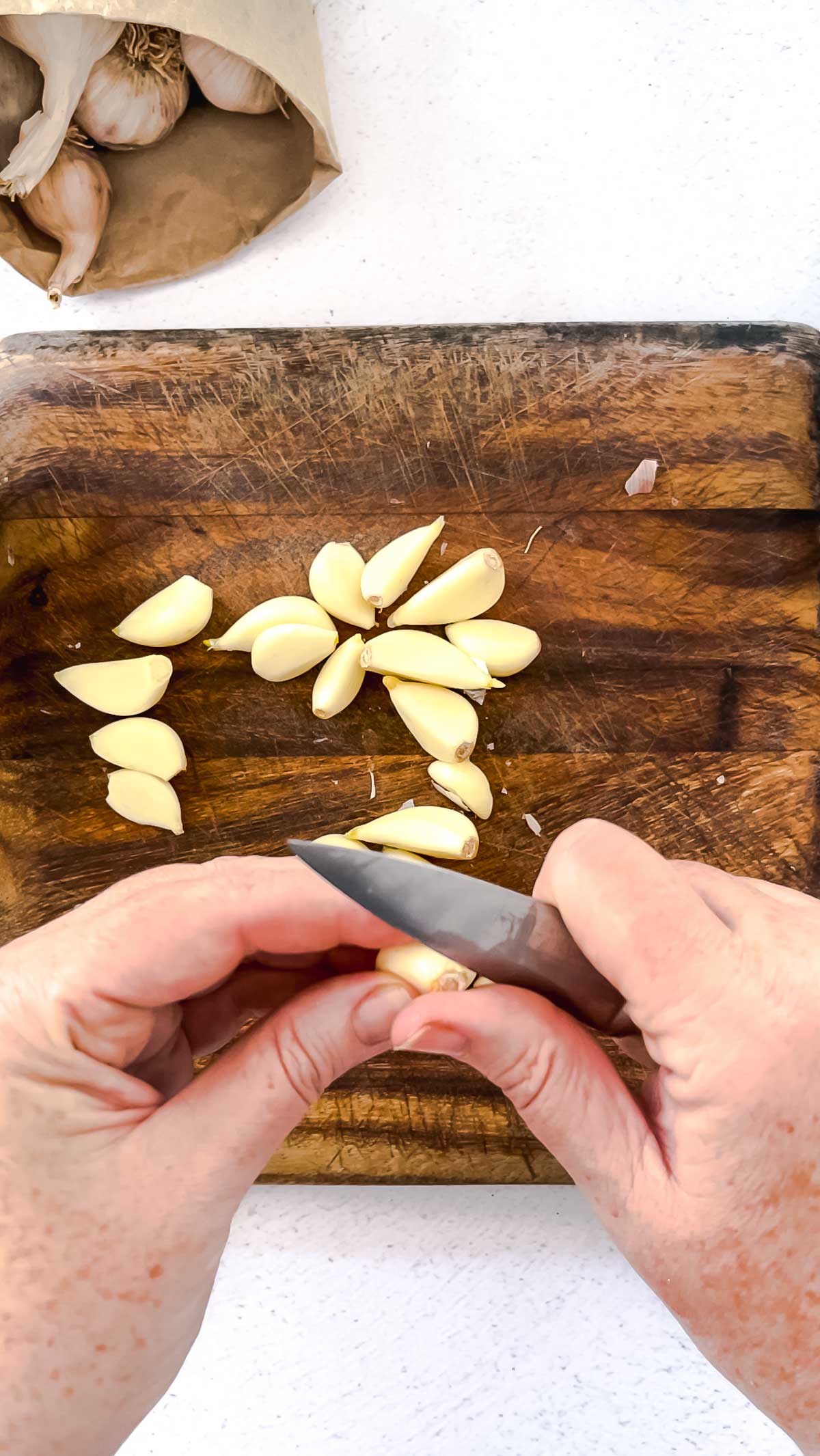 Two white hands are cutting the base off garlic cloves over a wooden chopping board.