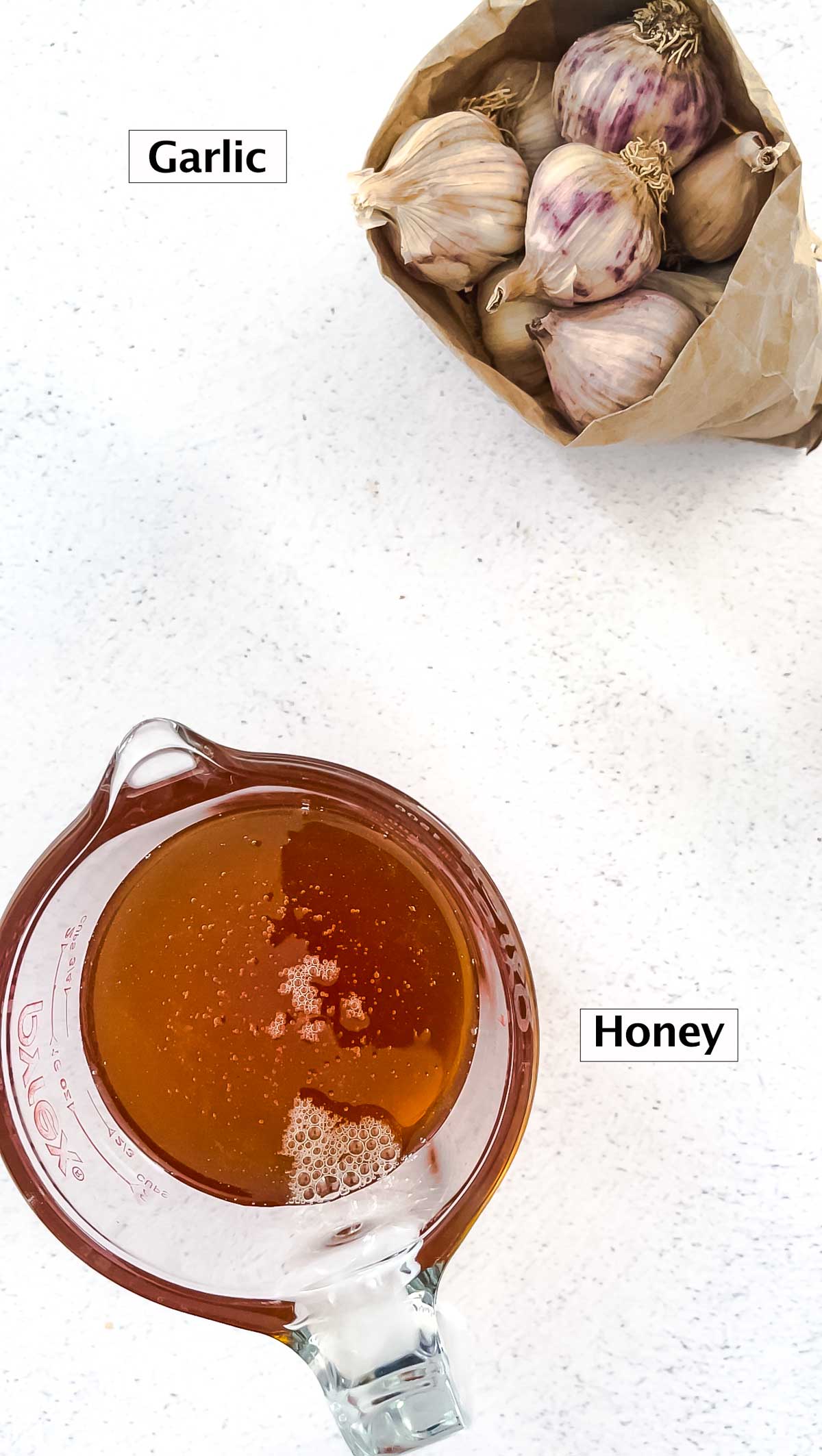 A brown bag filled with heads of garlic and a jug of honey are sitting on a kitchen bench.