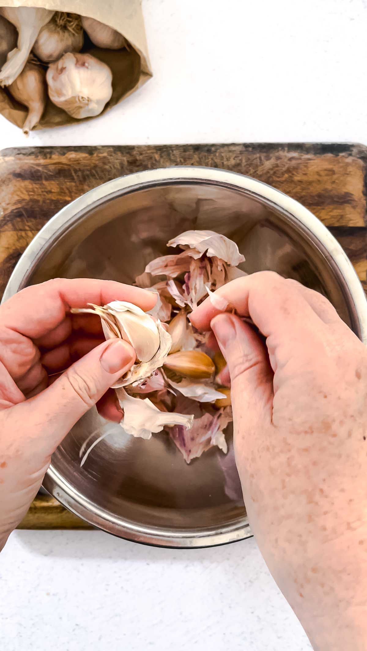 Two white hands are peeling garlic over a silver bowl. A brown paper bag filled with heads of garlic is also on the table.