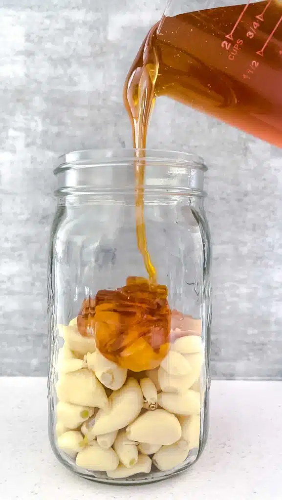 A large jar filled with peeled garlic cloves is standing on the bench. Honey is being poured into the jar to cover the cloves.