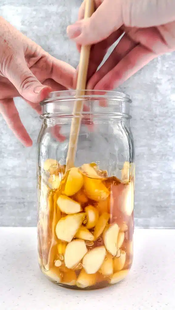 One white hand is holding steady a large jar filled with peeled garlic cloves and honey. Another white hand is using a chopstick to stir the garlic in the honey.