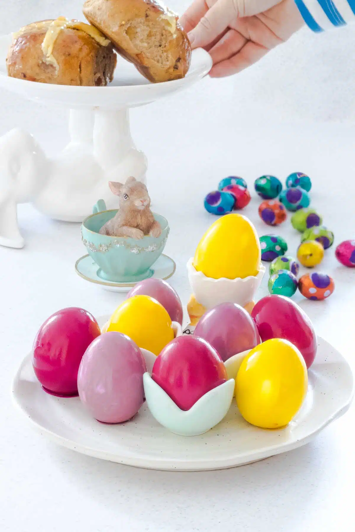 Brightly coloured pickled eggs are sitting on a white plate with some in egg cups. Behind them chocolate eggs are spread across the table, along with a bunny in a teacup decoration and a rabbit cake stand with hot cross buns on it.
