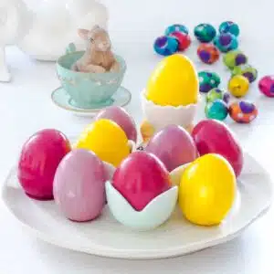 Various coloured, pickled eggs are sitting on a white plate, some in egg cups. They are yellow, pink, and purple. A few mini chocolate easter eggs have been scattered on the table behind the dish.