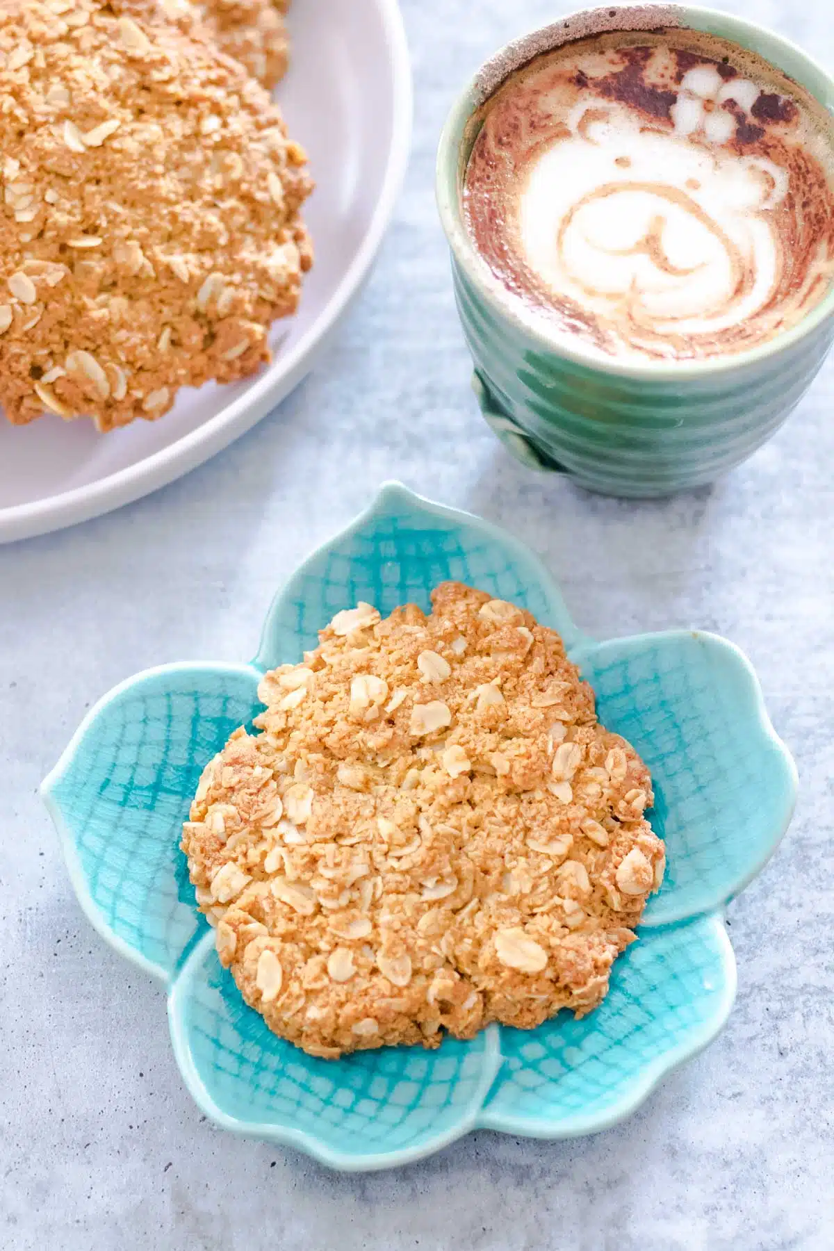 One Anzac biscuit is sitting on a small teal plate int he shape of a flower. A cup up coffee with bear latte art is sitting in the background. Beside it is a cooling rack with more biscuits.