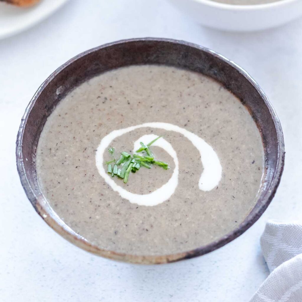 A small brown bowl is filled with mushroom soup and has a swirl of coconut cream and some chopped chives on top.