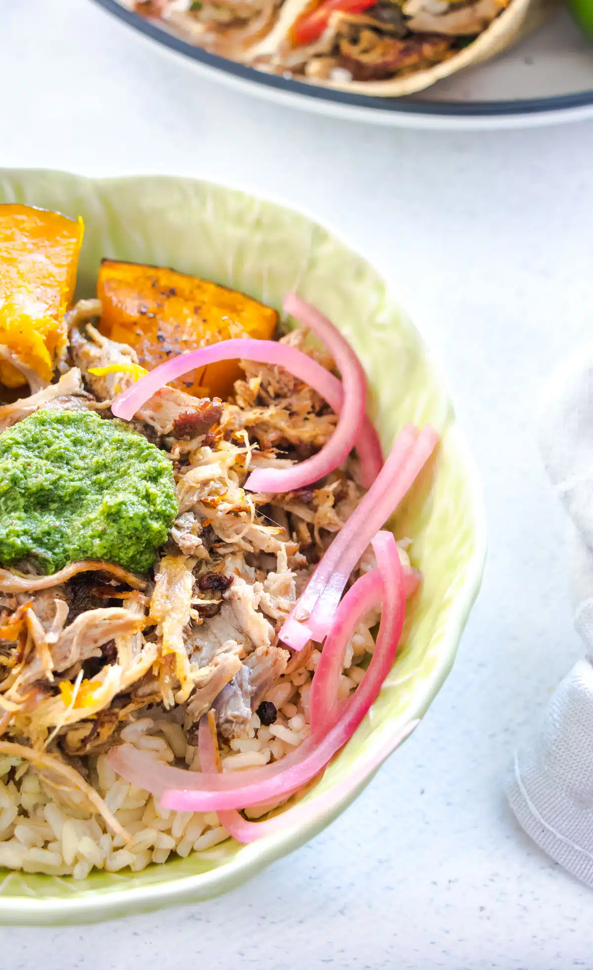 A green bowl filled with brown rice, topped with carnitas, baked veg, chimichurri, and pickled onions.