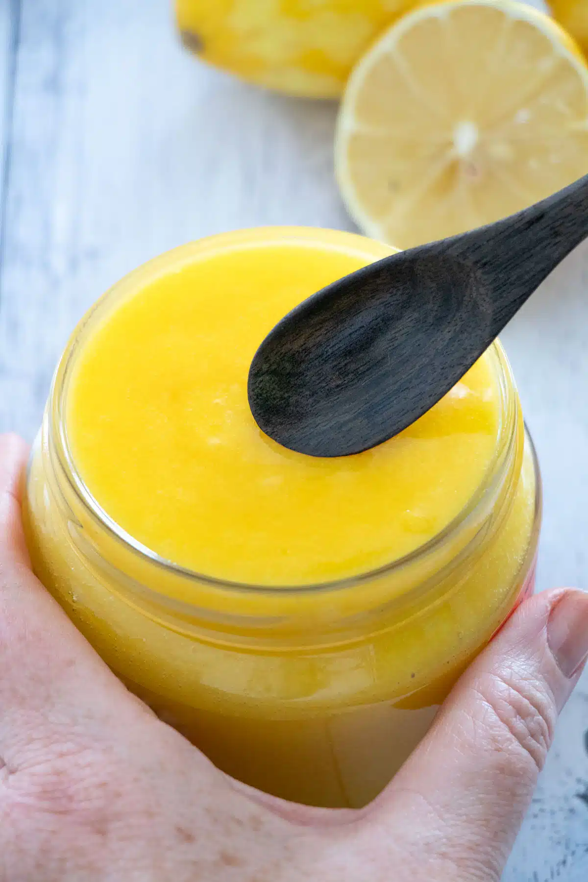 A white hand is holding a small jar of bright yellow, dairy-free lemon curd. A small dark-brown wooden spoon is about to be dipped into it.
