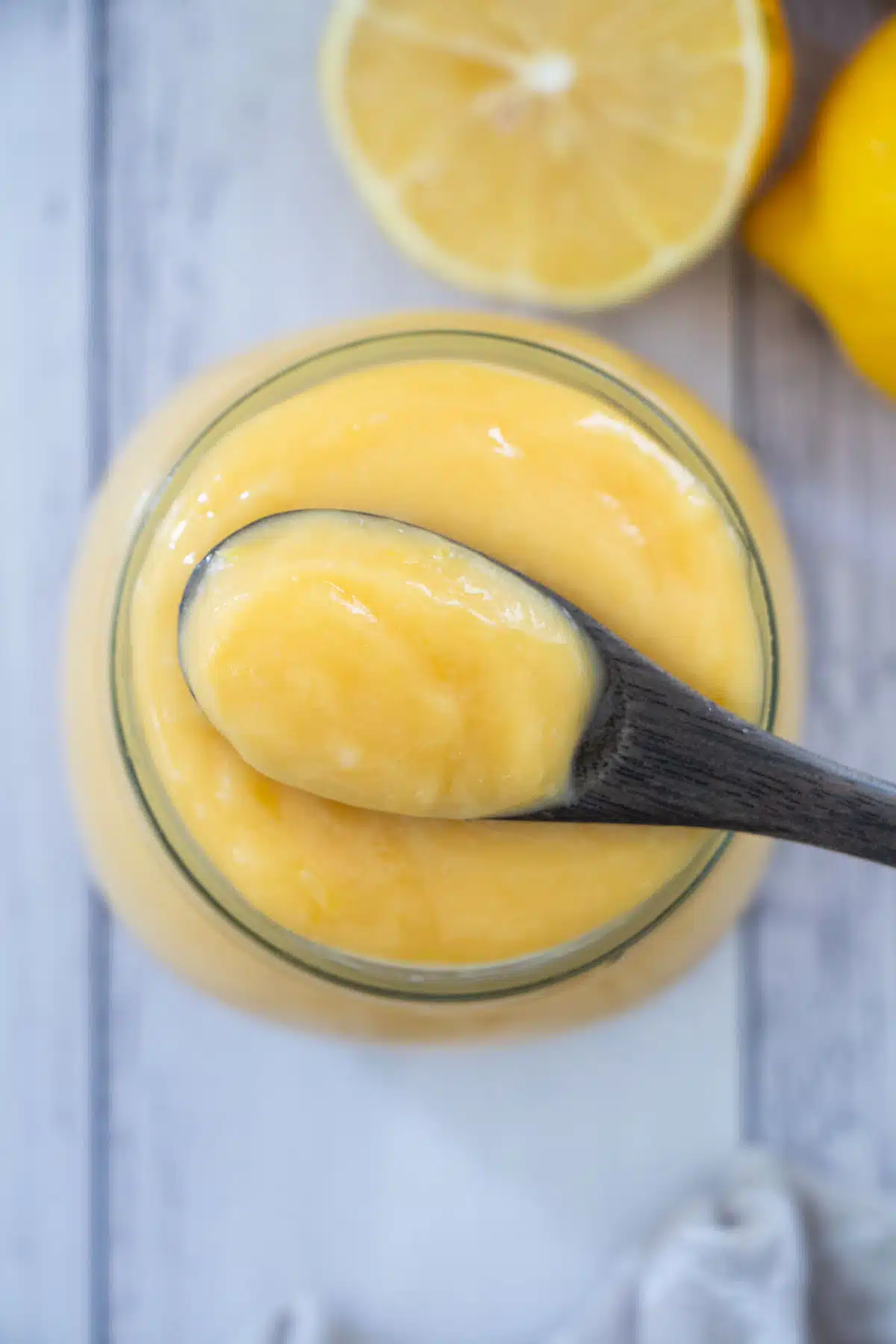 A close up looking at a jar of lemon curd with a small wooden spoon on top that has been dipped into the curd. A few lemons can be seen in the background.