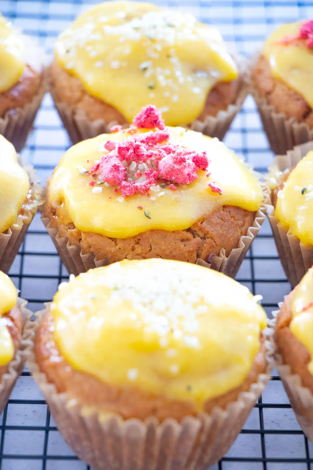 Nine lemon curd muffins have been laid out on a black cooling rack. Each have been topped with extra bright yellow lemon curd and sprinkled with freeze dried strawberries or hemp seeds.