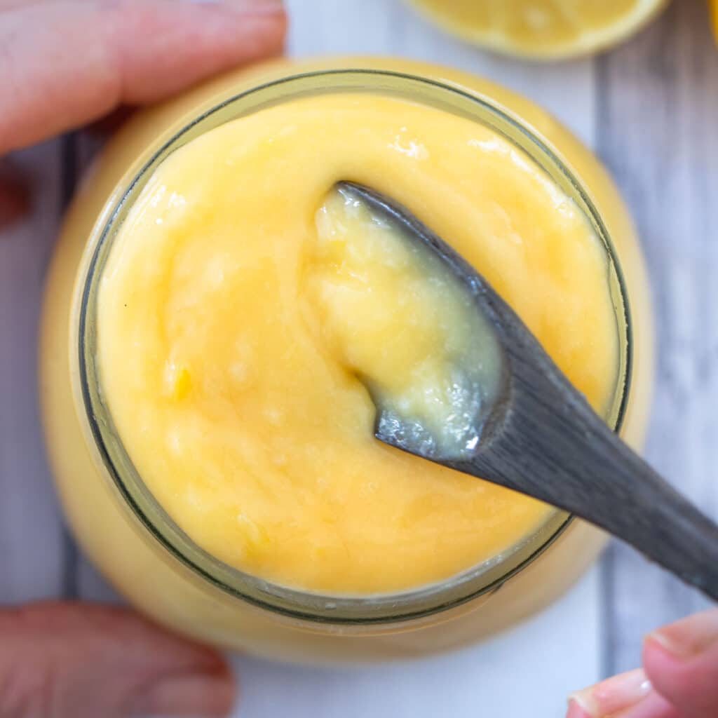 A jar of lemon curd is being held by a white hand. A small dark-brown wooden spoon is being dipped into the jar to scoop some up.