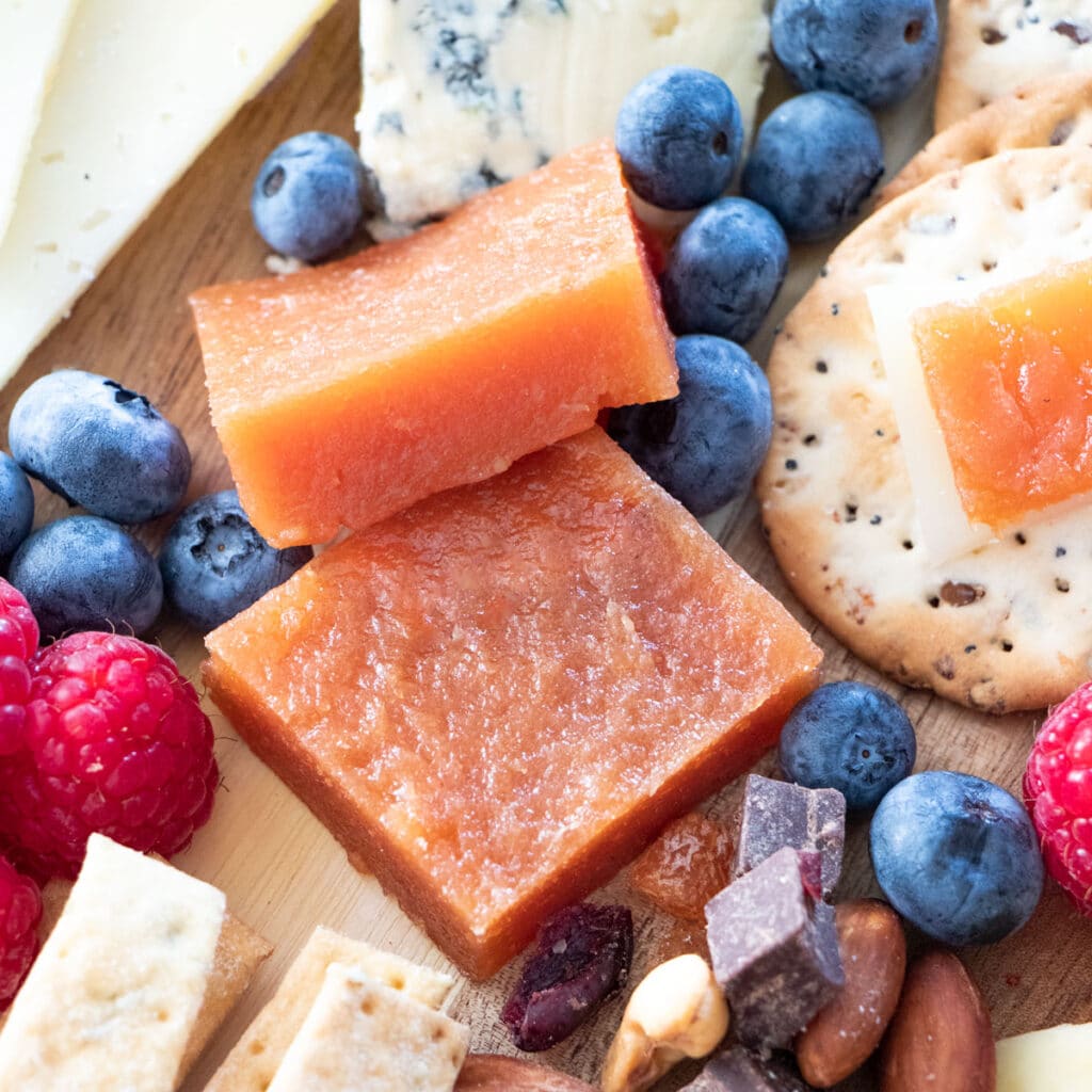 A square image showing a close up of deep pink quince paste cut into squares and sitting on a cheese board. It's surrounded by cheese, crackers, nuts and dried fruit, as well as berries.