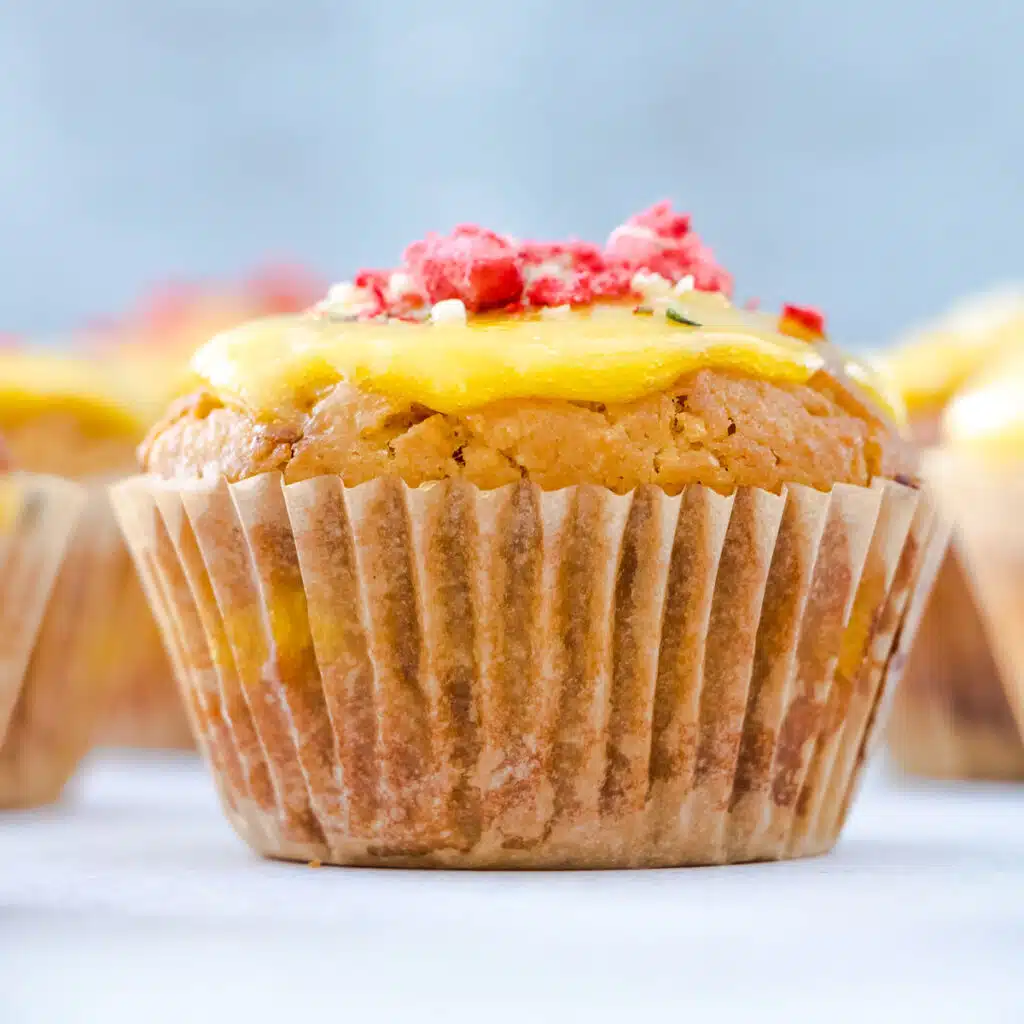 A square image of a single lemon curd muffin topped with more curd and freeze dried strawberries. Other muffins are surrounding it on the edges of the table but these are out of focus.