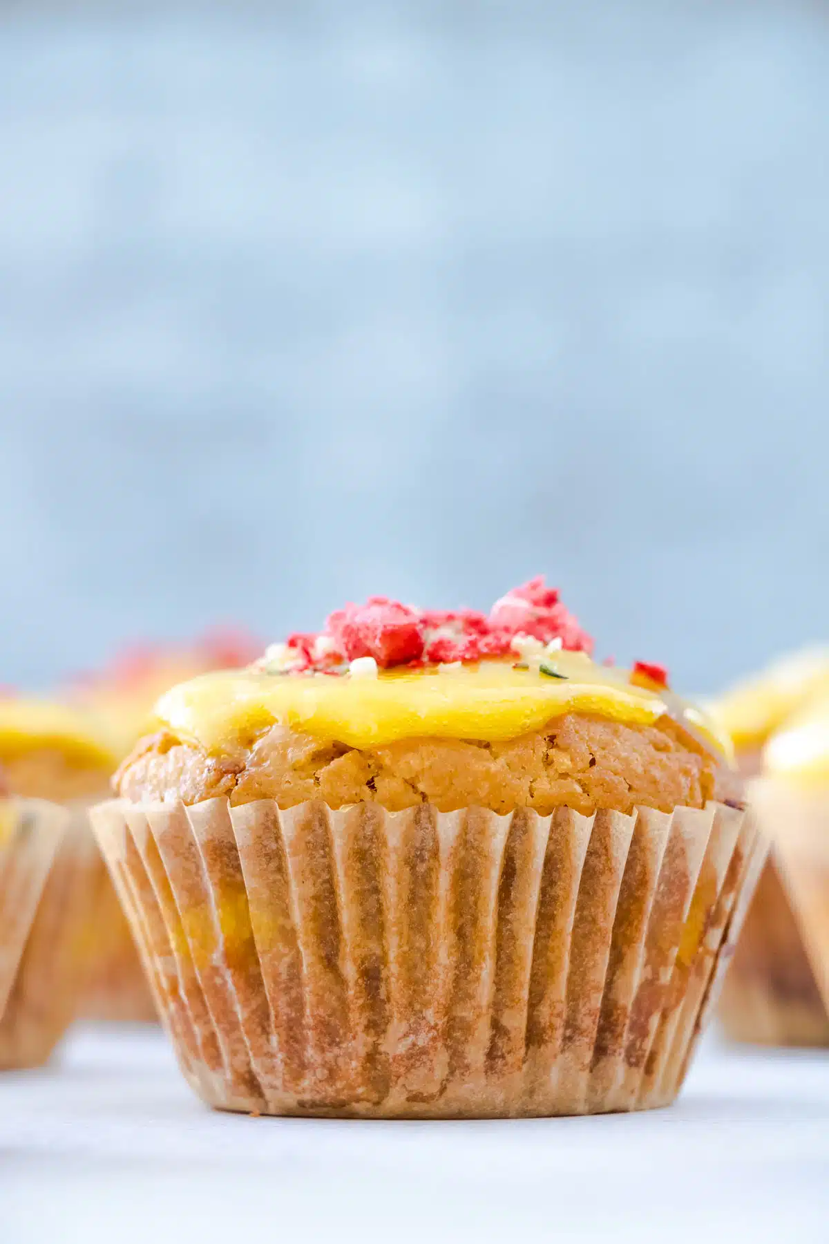 A lemon curd muffin topped with more curd and freeze dried strawberries is at the centre of the image. Other muffins are surrounding it on the table but these are out of focus.