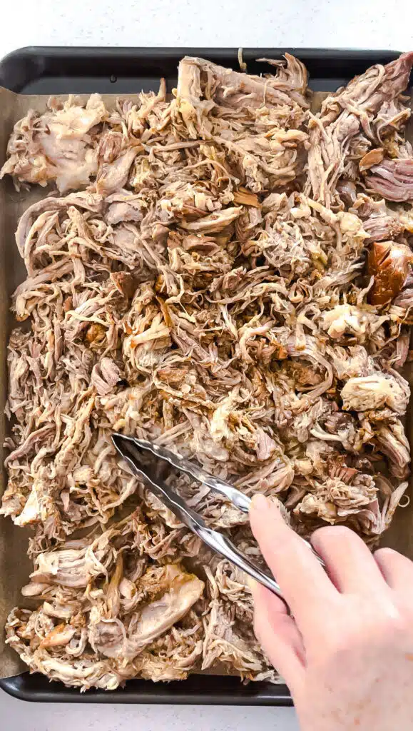 A white hand is spreading out the shredded pork meat on a tray lined with baking paper.
