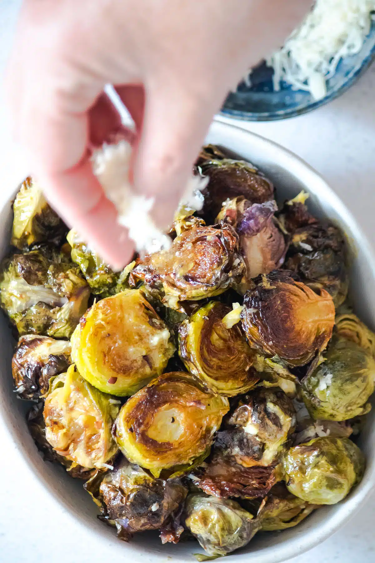 A white hand is sprinkling grated parmesan cheese over a bowl of caramelized Brussels sprouts.
