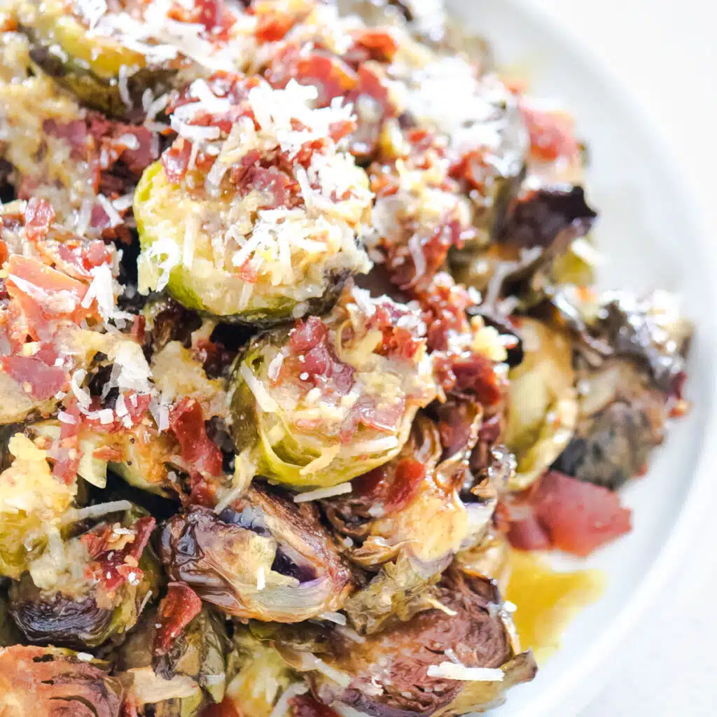 A close-up of roasted Brussels sprouts piled on a plate and covered cooked prosciutto and grated parmesan cheese.