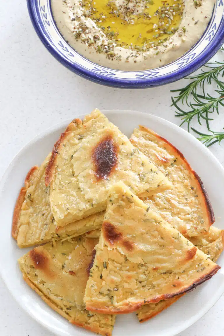 A plate of triangular-shaped socca slices have been served beside a bowl of hummus. Some sprigs of rosemary are also on the table.