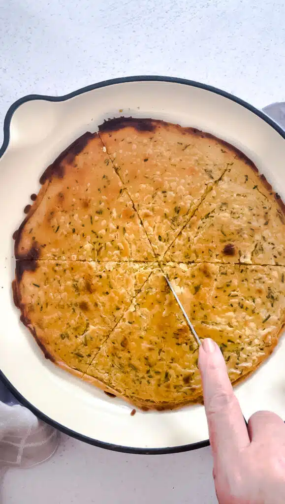 A white hand is cutting the cooked chickpea pancake into triangular slices.