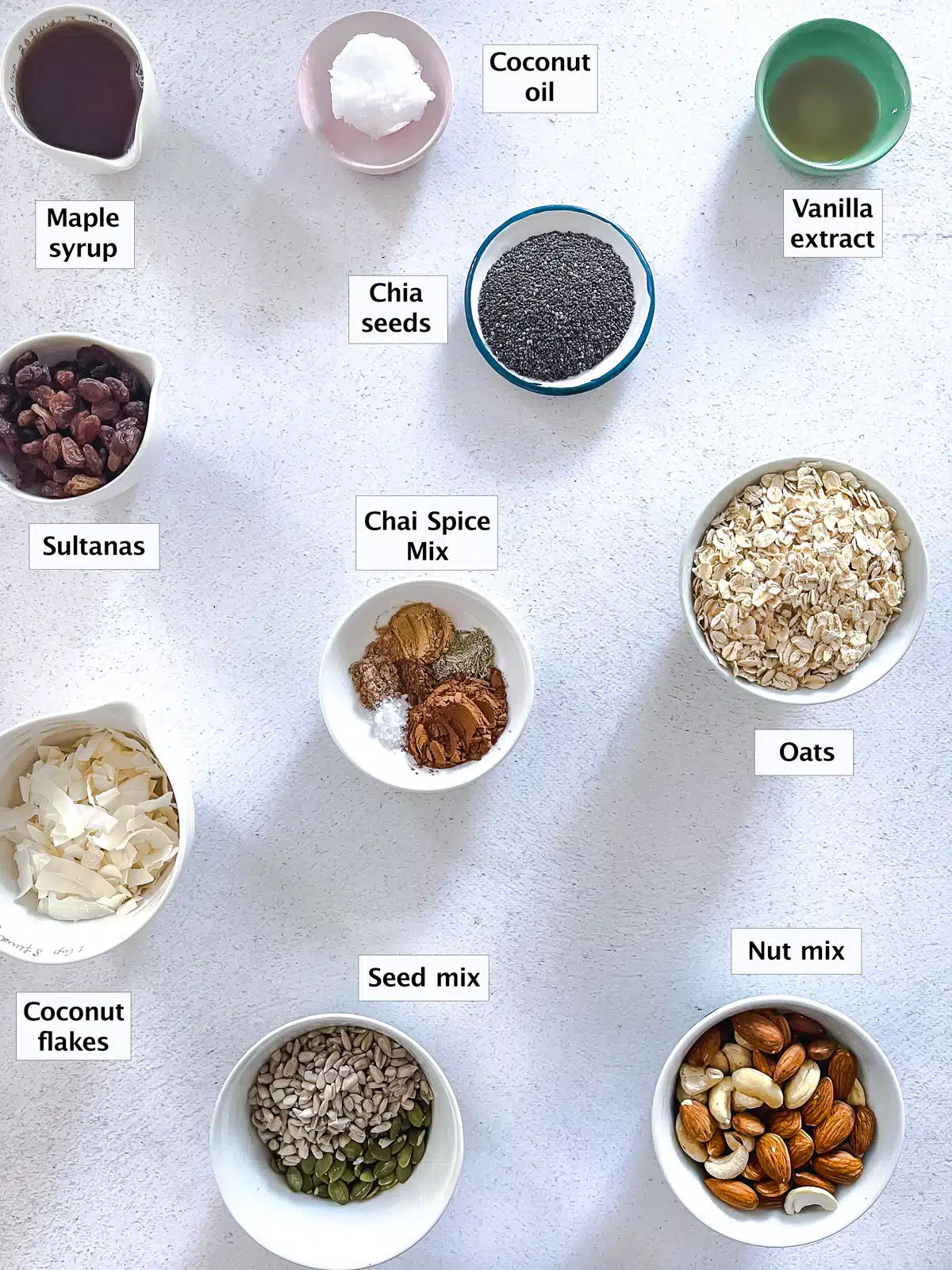 All the ingredients needed to make chai spiced granola have been laid out in small bowl across a white bench.