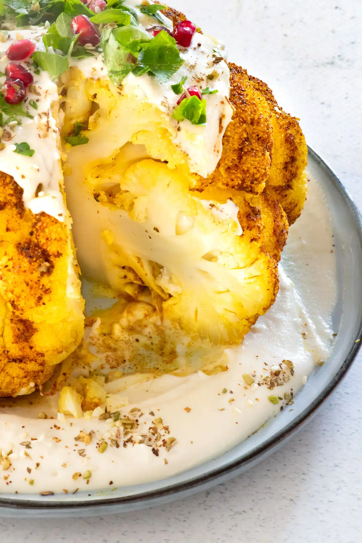 A close up looking into the cut roasted cauliflower. It's sitting on tahini, which is on a blue plate with a black. It's also been drizzled with tahini and covered in chopped parsley, pomegranate, and dukkha.