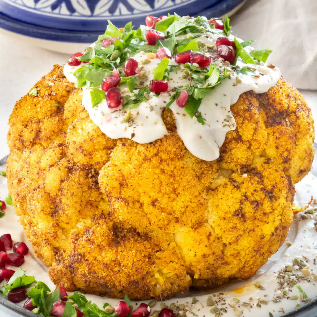 A square image showing a close up of a whole roasted cauliflower sitting on a plate of tahini. It's also topped with tahini and has been sprinkled with chopped parsley, pomegranate arils and dukkha.