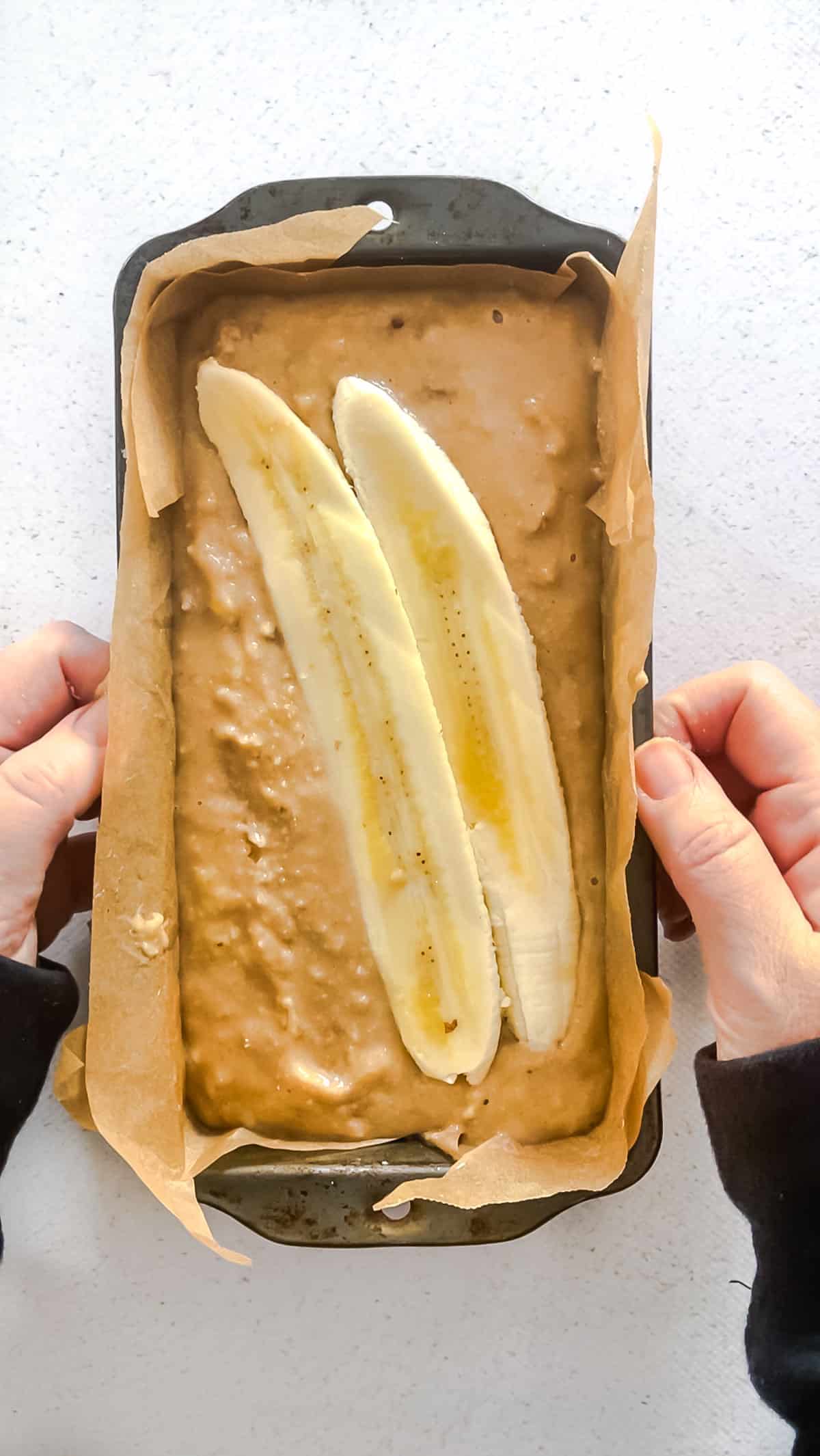 Two white hands are holding a loaf tin lined with brown baking paper. It's been filled with tahini banana bread batter and topped with two slices of banana.