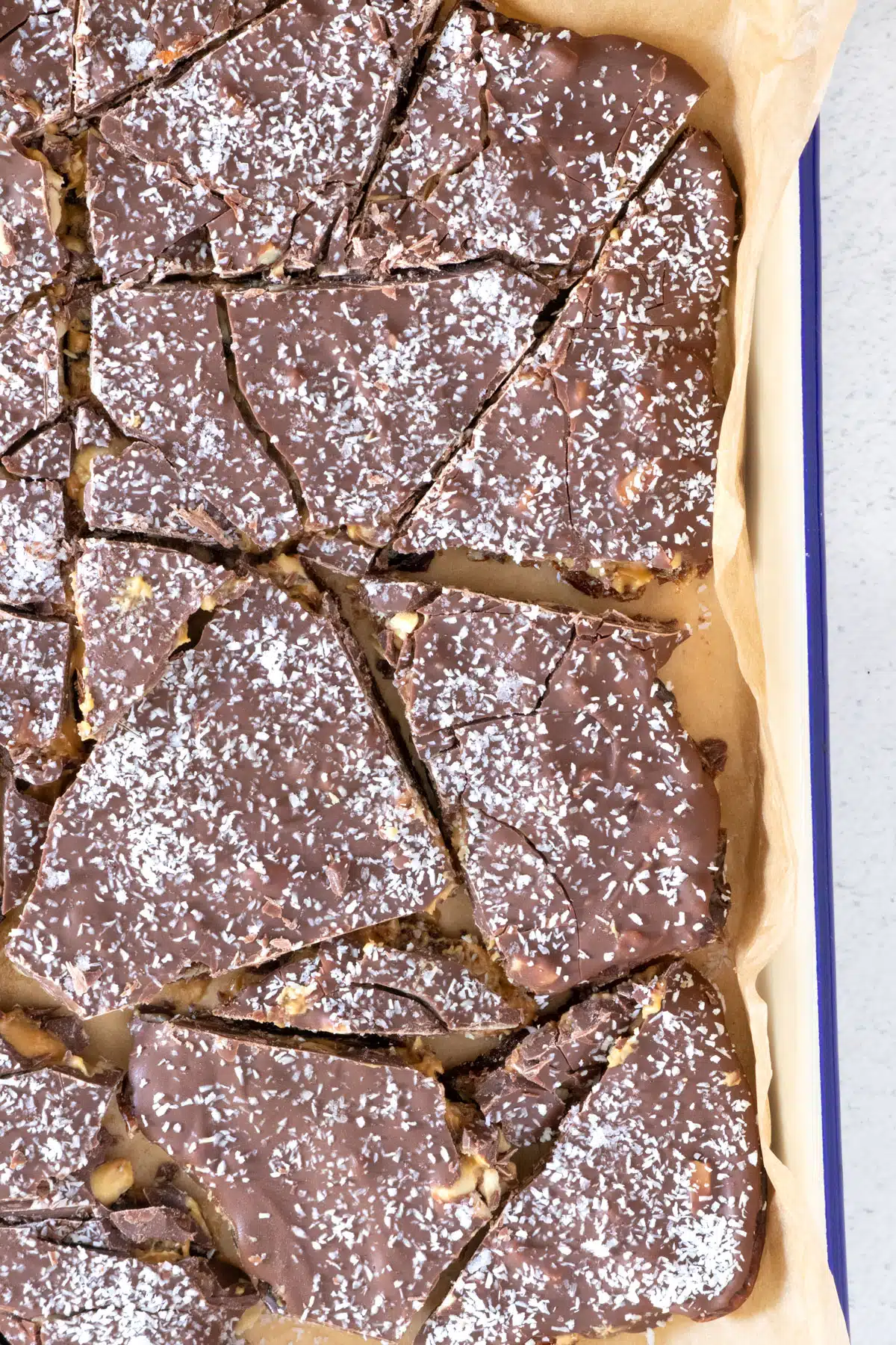 Easy Chocolate date bark has been chopped up in various shapes and is sitting on brown baking paper on a white tray with blue trim. The top has been sprinkled with coconut.