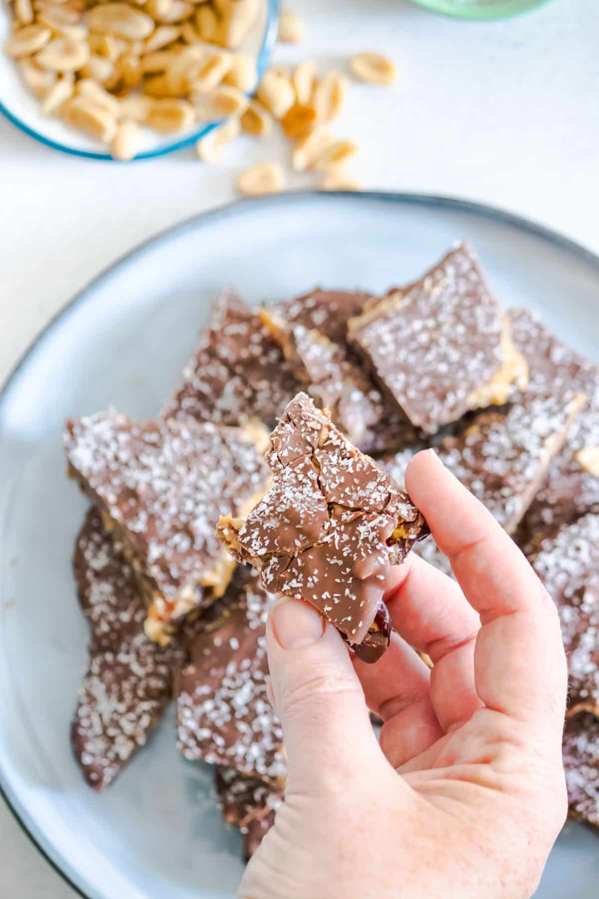 A white hand is holding up a piece of chocolate date bark. Underneath a blue plate is filled with more date bark and a bowl of peanuts and coconut can just be seen in the frame.
