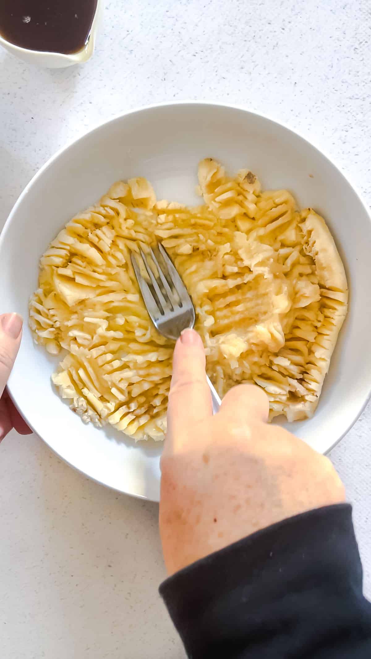 A white hand is pushing a fork into a bowl full of bananas to mash them.
