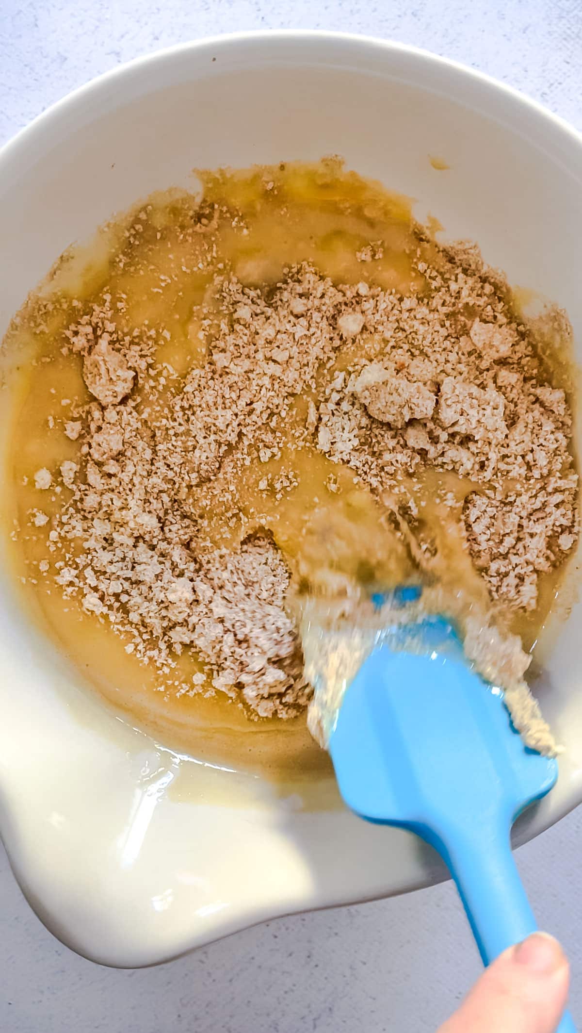 A white hand is holding a blue spatula that is stirring together the wet and dry ingredients for the tahini banana bread.