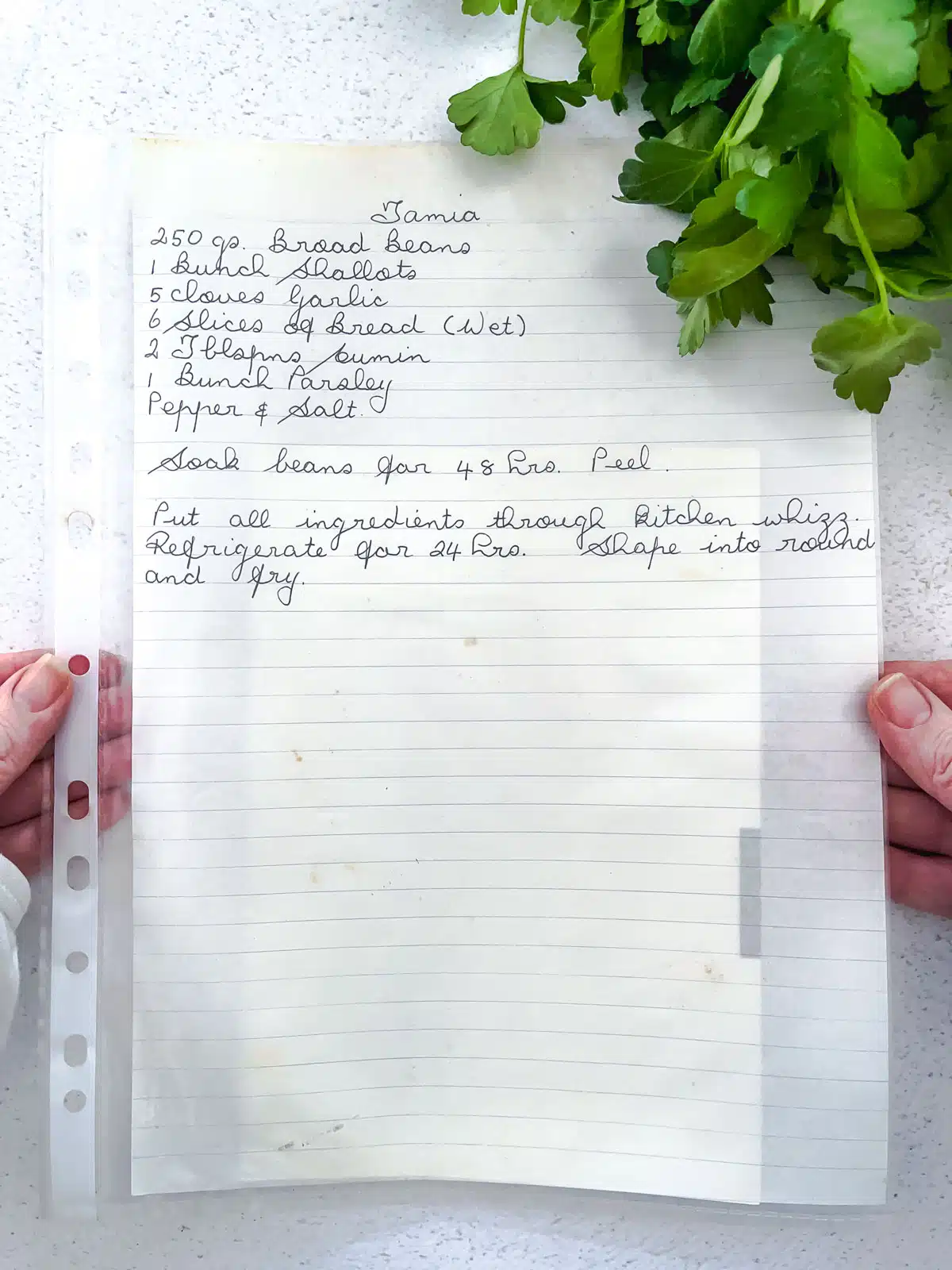 2 white hands are holding a lined piece of paper with a recipe written on it. Some bright green parsley is on the bench at the top right of the image.