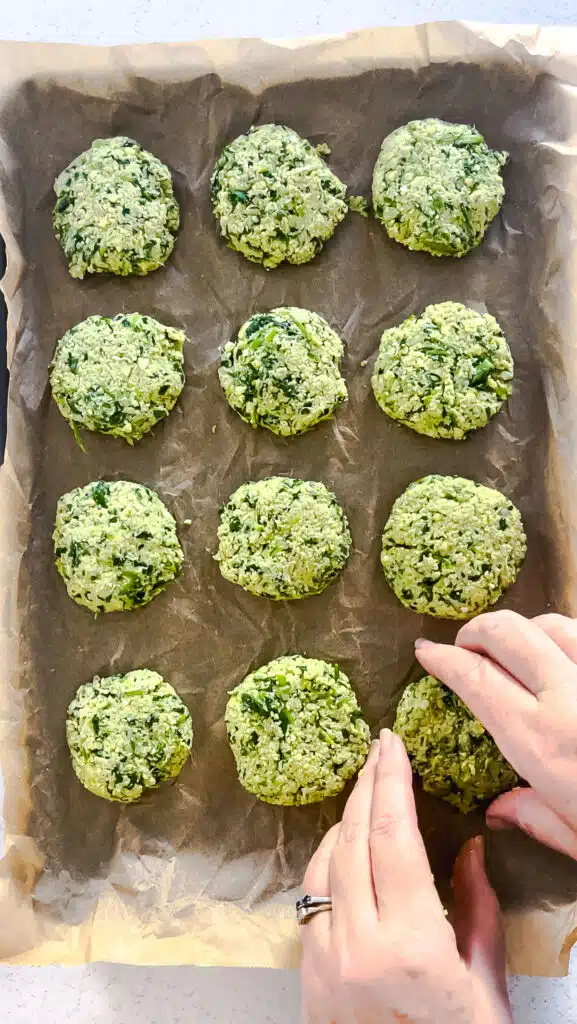 11 green falafel have been placed on a lined baking tray. Two white hands are placing the 12th down.