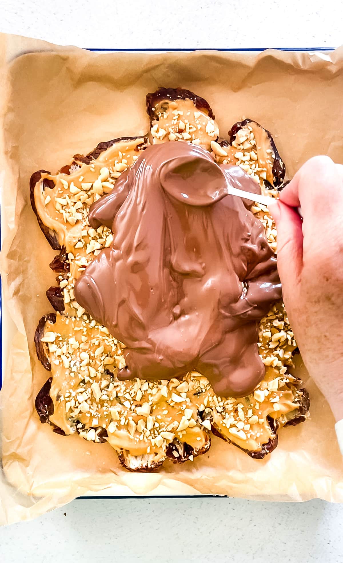 A white hand is spreading melted chocolate over the top of the date bark with the back of a spoon.