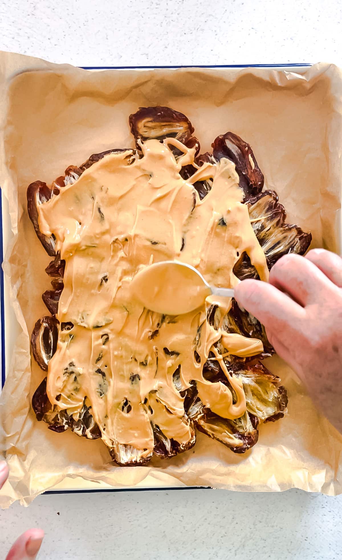 A white hand is holding a spoon that is spreading peanut butter over dates flattened out on a baking tray lined with baking paper.
