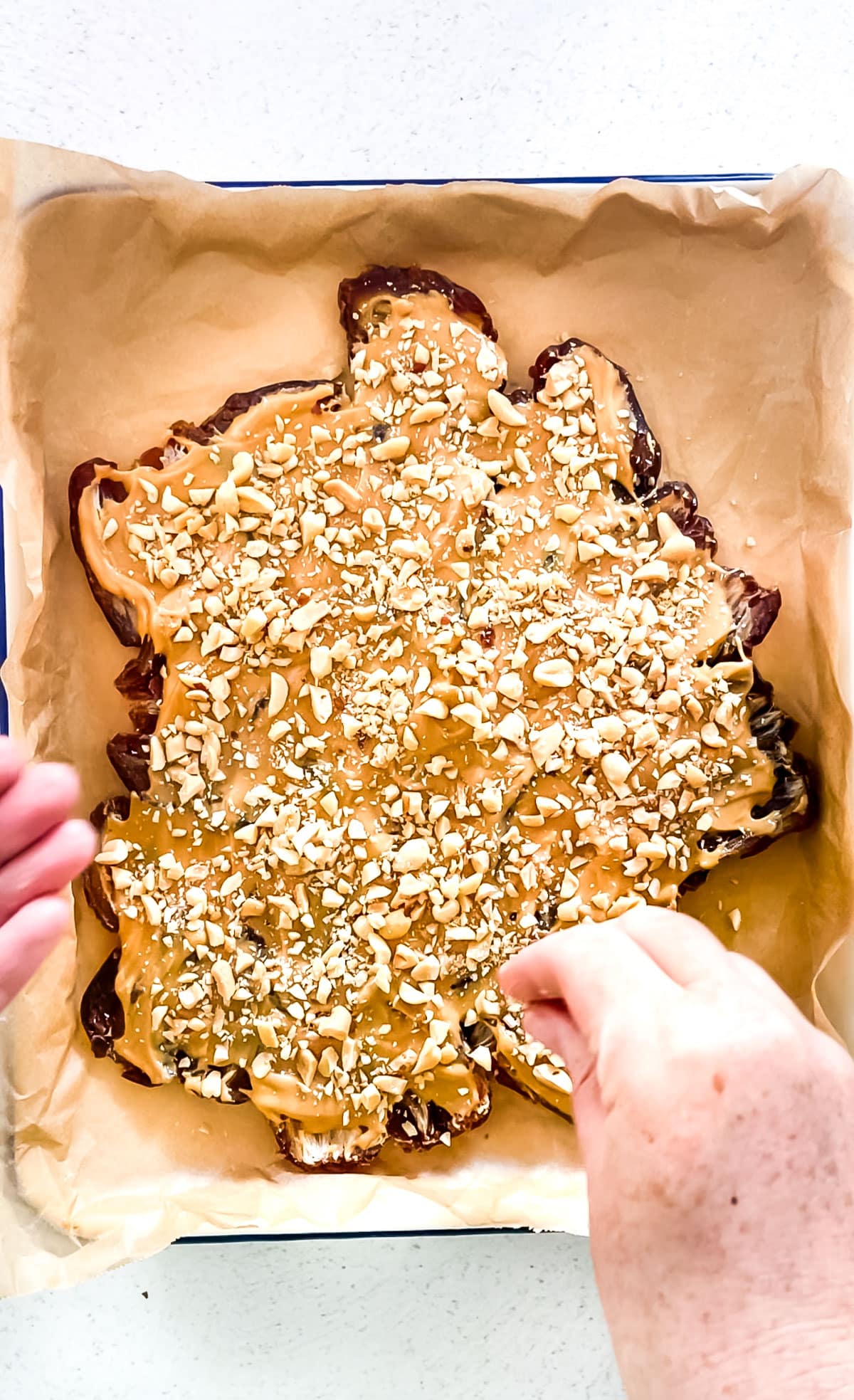 A white hand is sprinkling chopped peanuts over the top of the date bark. They're falling onto peanut butter spread over the dates.
