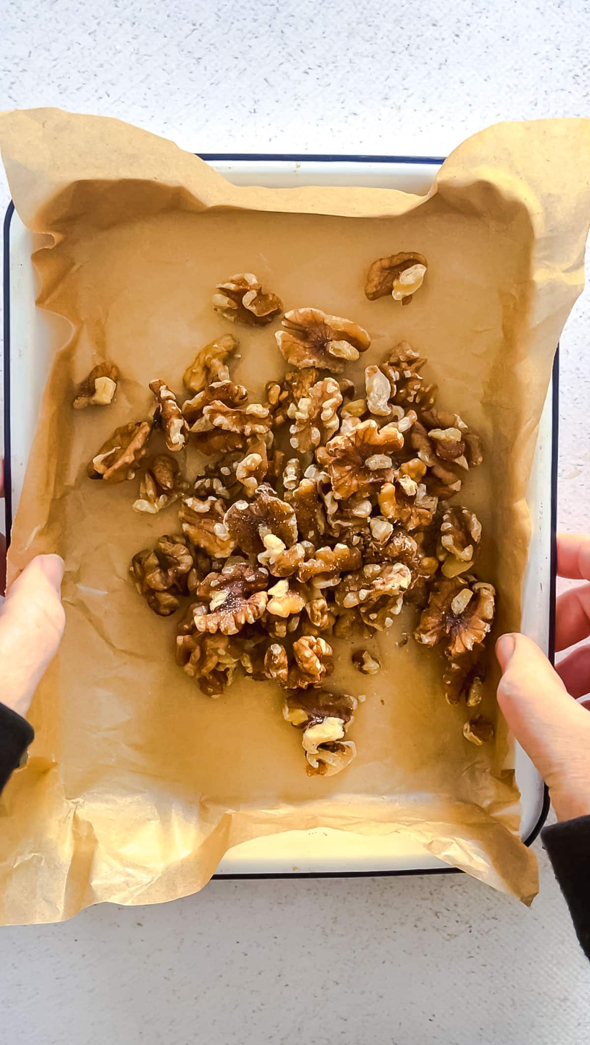 Walnuts are scatter over a white tray lined with brown baking paper. Two white hands are holding the edges of the tray.