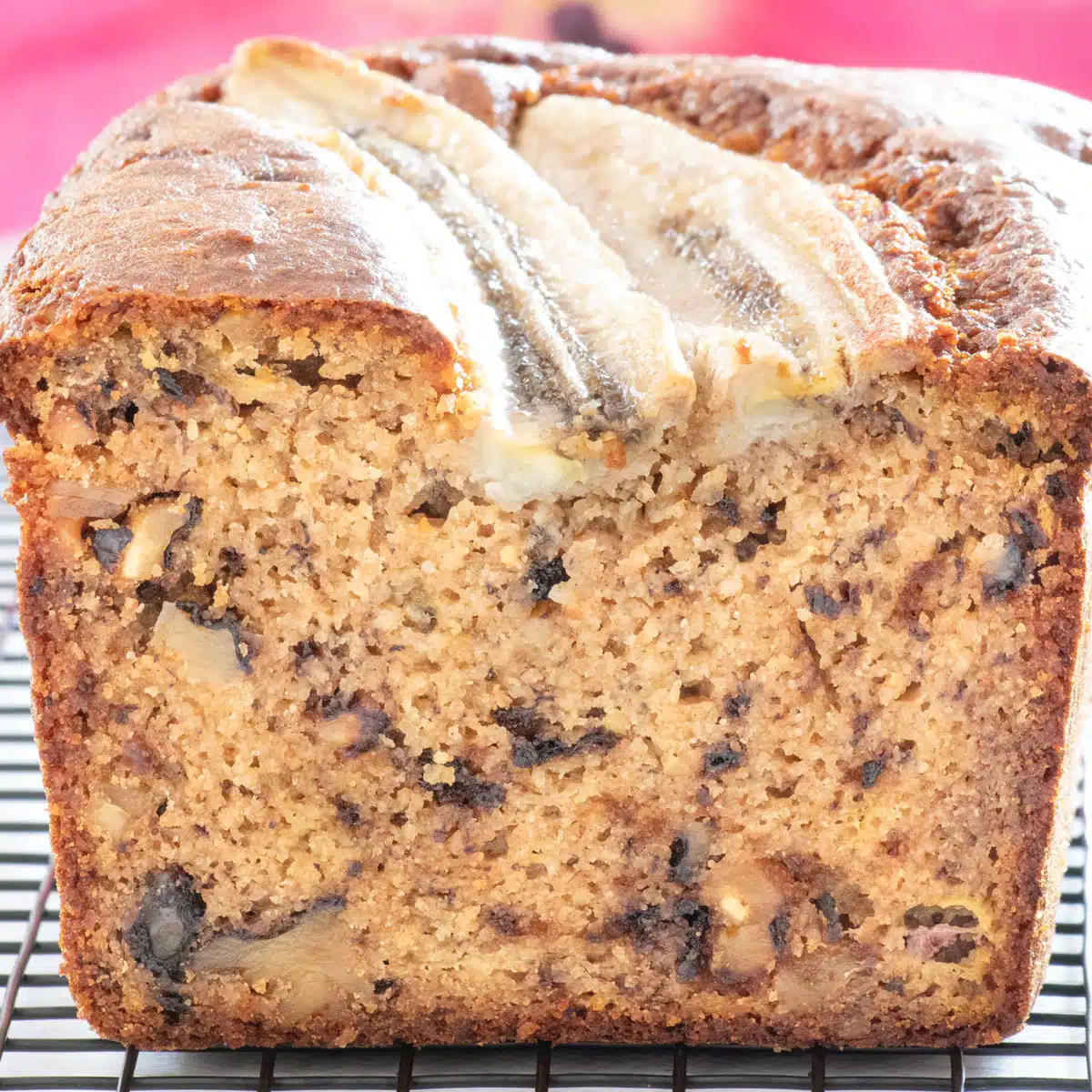 A square image showing a cut loaf of banana bread front on. A pink flower fills the background.
