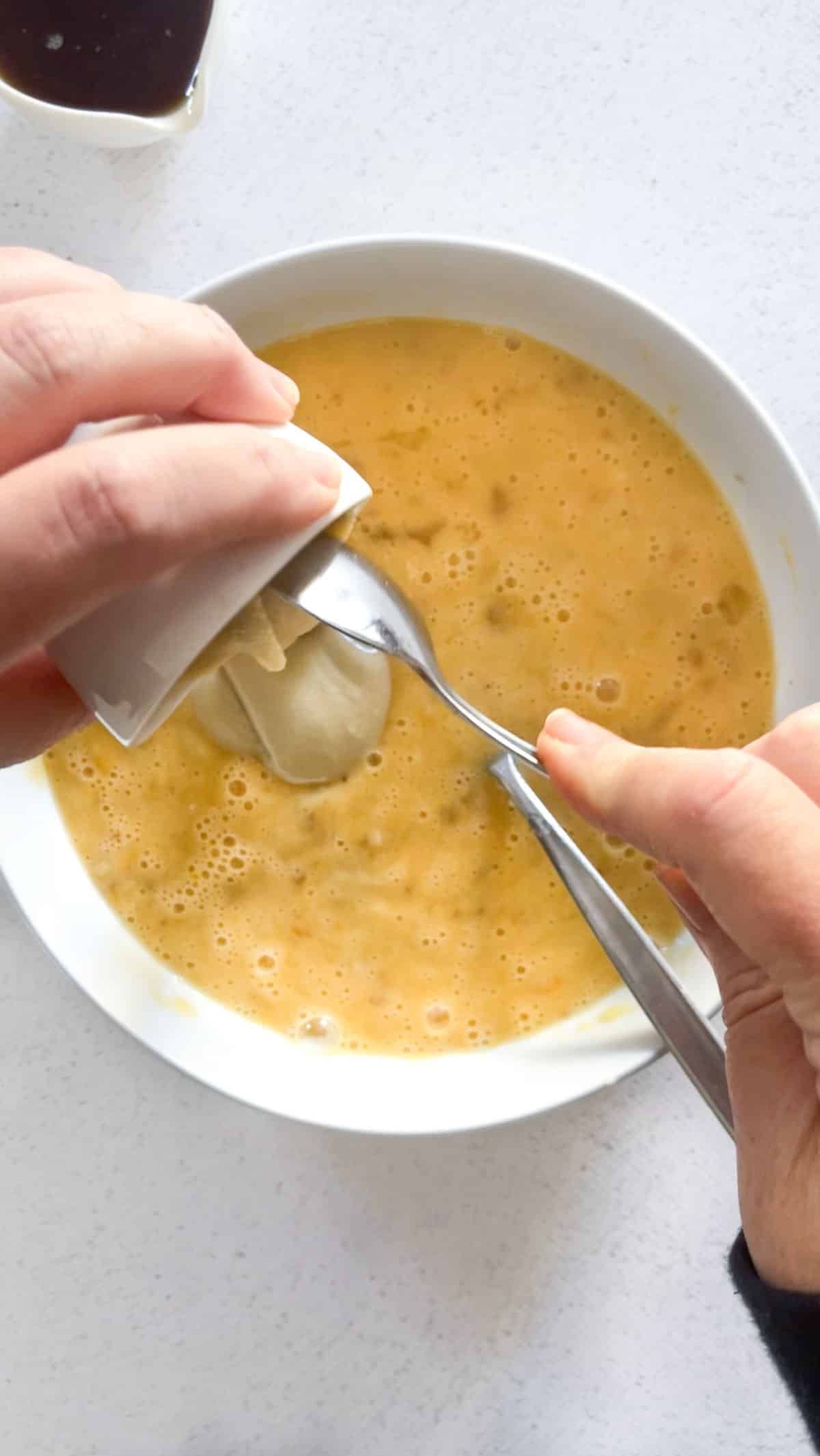 Two white hands can be seen hovering above a white bowl with tahini banana bread batter. One is holding a small bowl of tahini, which is being poured out, the other is holding a spoon that is scraping the tahini out into the bowl below.
