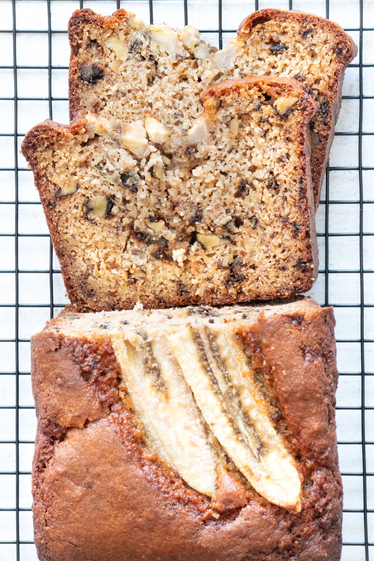 Look down onto a golden brown loaf of Tahini Banana Bread with two slices of banana on top. The loaf is sitting on a black, wire cooling rack and has been sliced. Two slices have fallen in front of it onto the rack.
