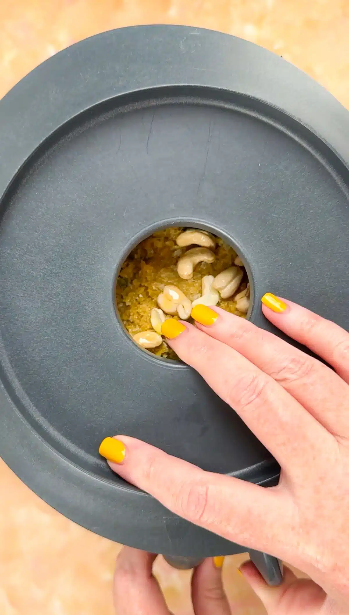 A white hand with yellow painted nails is putting the lid on a food processor. A small hole in the top of the lid shows the ingredients inside.