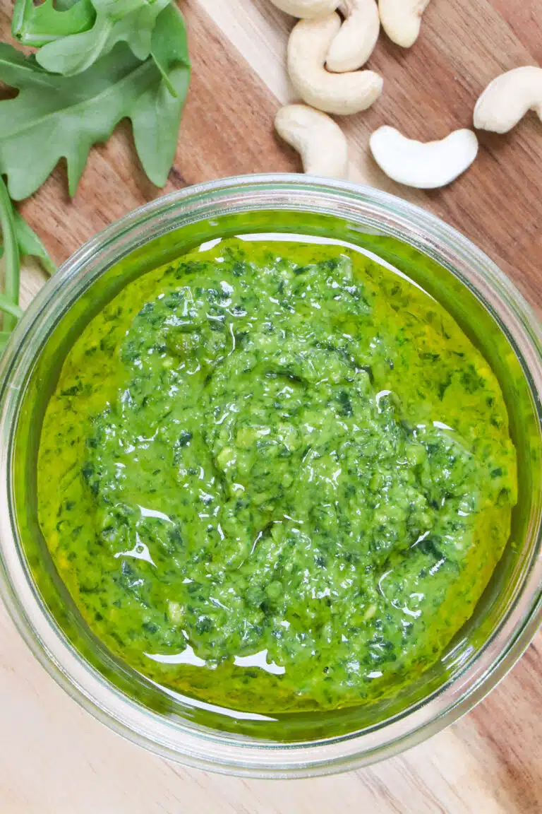 Looking down into a jar of bright green rocket (arugula) pesto that's sitting on a wooden board. Some rocket and cashews are scatted on the table beside the jar.