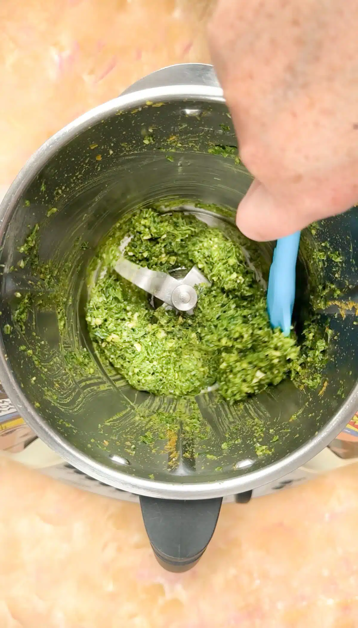 A white hand is scraping down the inside of a food processor with a blue spatula to get all the pesto off the sides.