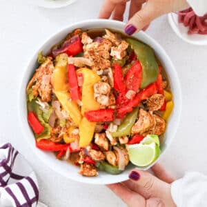 A close up of a bowl filled with chiacken fajitas being held by two white hands.