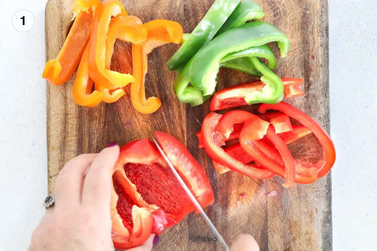 Red, green, and orange bell peppers are on a wooden chopping board. The red pepper is being cut with a large knife.