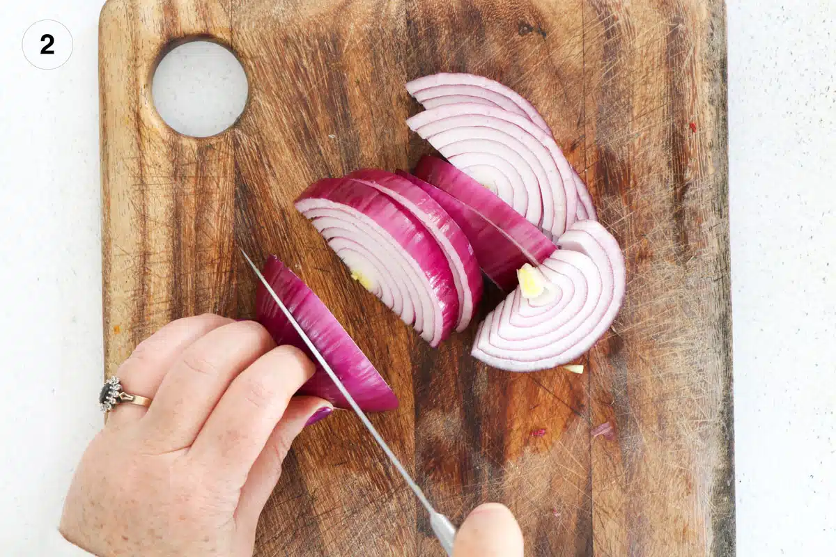 A red onion is cut in half and sitting on a wooden chopping board. One half is being held and sliced.