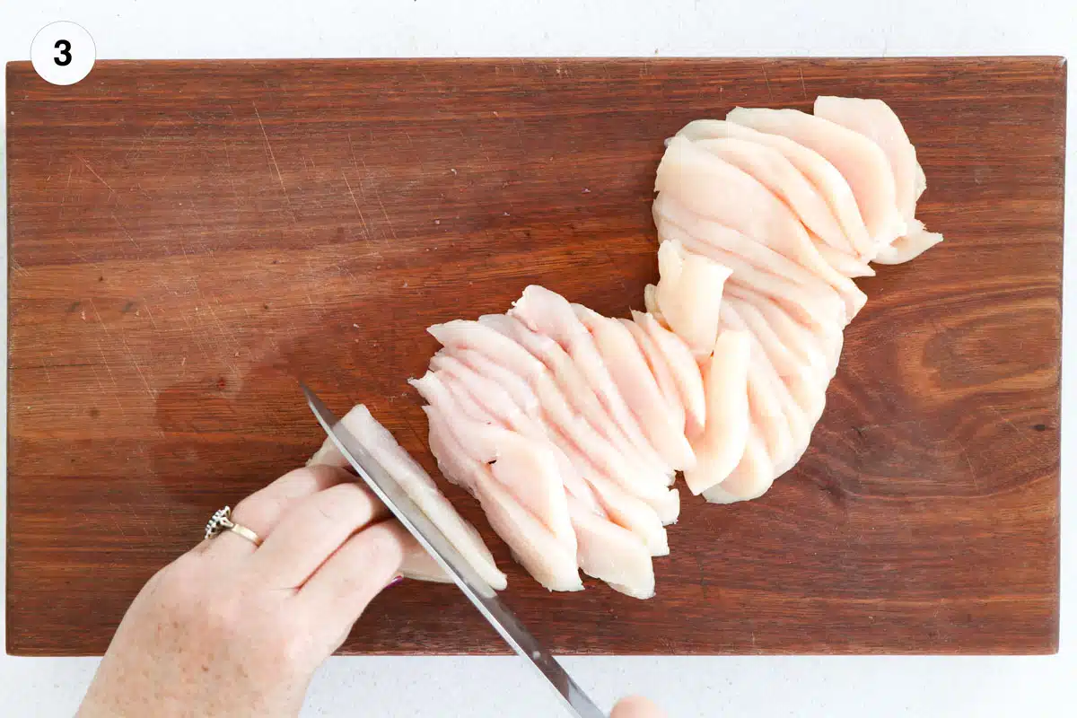 A chicken breast is being sliced with a large silver knife on a wooden chopping board.